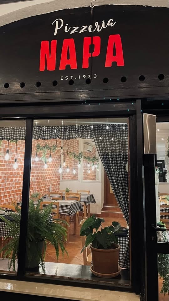 Vhod v Pizzerio Napa zvečer: črn napis z rdečim NAPA, za steklom opečnat zid, karirasti prti in žarnice.